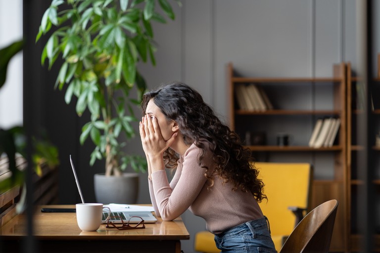 Enttäuschte Büroangestellte, die mit einer Aufgabe kämpft, unproduktiv ist, müde und überarbeitet – eine weibliche Mitarbeiterin bedeckt ihr Gesicht mit den Händen und denkt über eine Lösung nach, während sie an einem Tisch mit einem Laptop sitzt. 