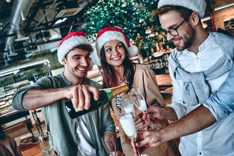 Weihnachtsfeier: Kolleginnen und Kollegen schütten sich Sekt in Gläser ein und tragen Nikolausmützen, im Hintergrund ist ein geschmückter Tannenbaum.