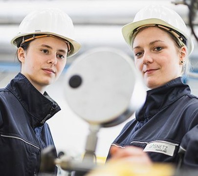 Zwei junge Frauen schauen auf ein weißes Gerät und tragen Helme.