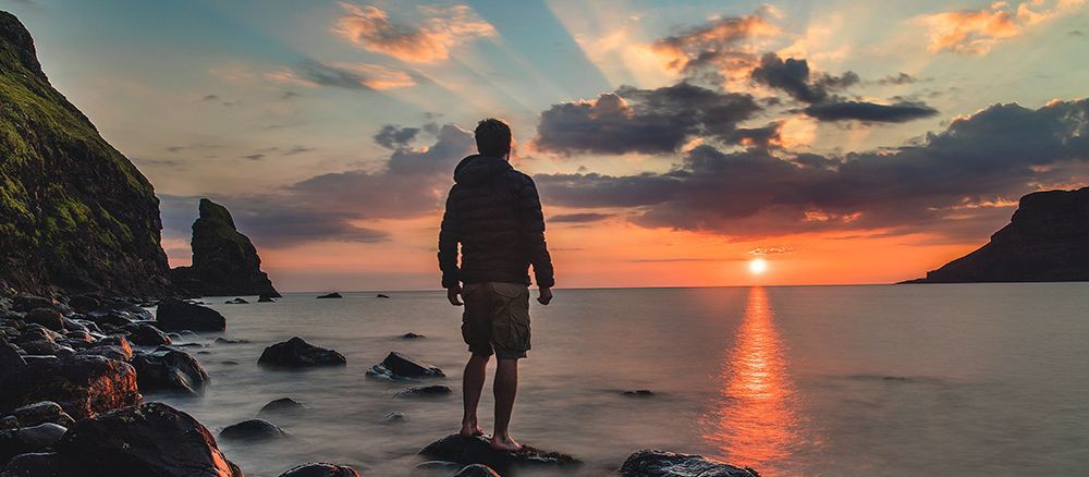 Junger Azubi steht im Ausland auf einem Stein im Meer und blickt auf Sonnenuntergang