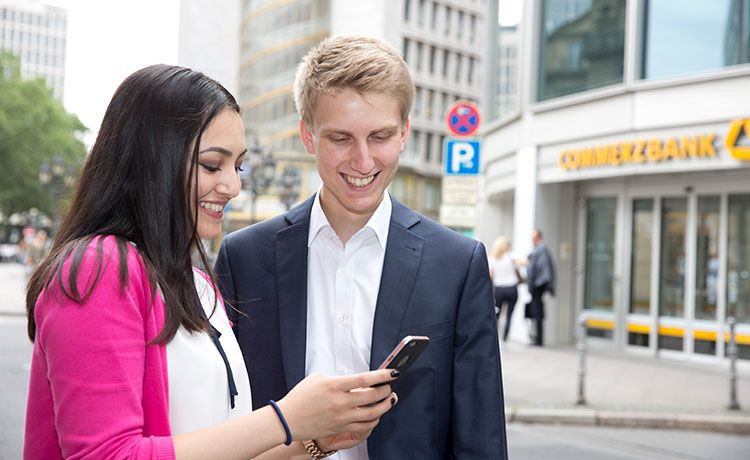 eine Frau und ein Mann stehen vor der Commerzbank und schauen auf ein Handy.