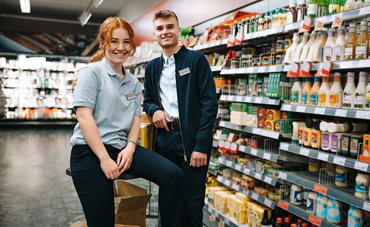 Zwei junge Handelsfachwirte stehen in einem Supermarkt vor einem Regal und lächeln in die Kamera.