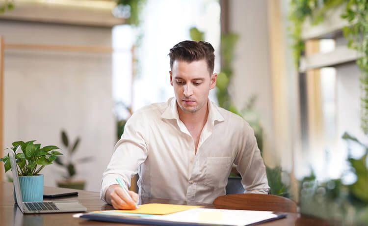 Junger Mann mit Hemd sitzt an einem Schreibtisch und macht sich Notizen.