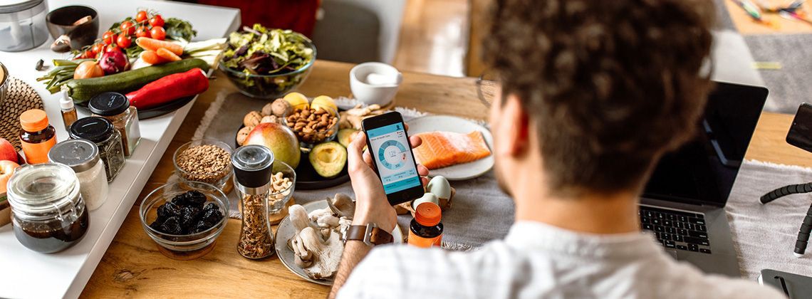 Junger Ernährungsberater sitzt an einem Tisch voller gesunder Lebensmittel und checkt eine Kalorientracker-App am Smartphone.