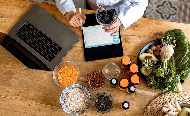 Blick von oben auf einen Tisch voller gesunder Lebensmittel, an dem ein Ernährungsberater sitzt, der an einem Tablet einen Ernährungsplan erstellt.