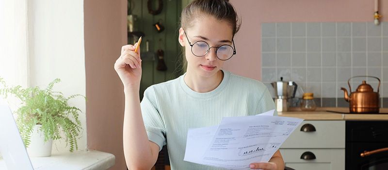 Junge Auszubildende mit Brille sitzt in der Küche, hält einen Stift in der Hand und schaut auf Dokumente, die sie in der Hand hält. 