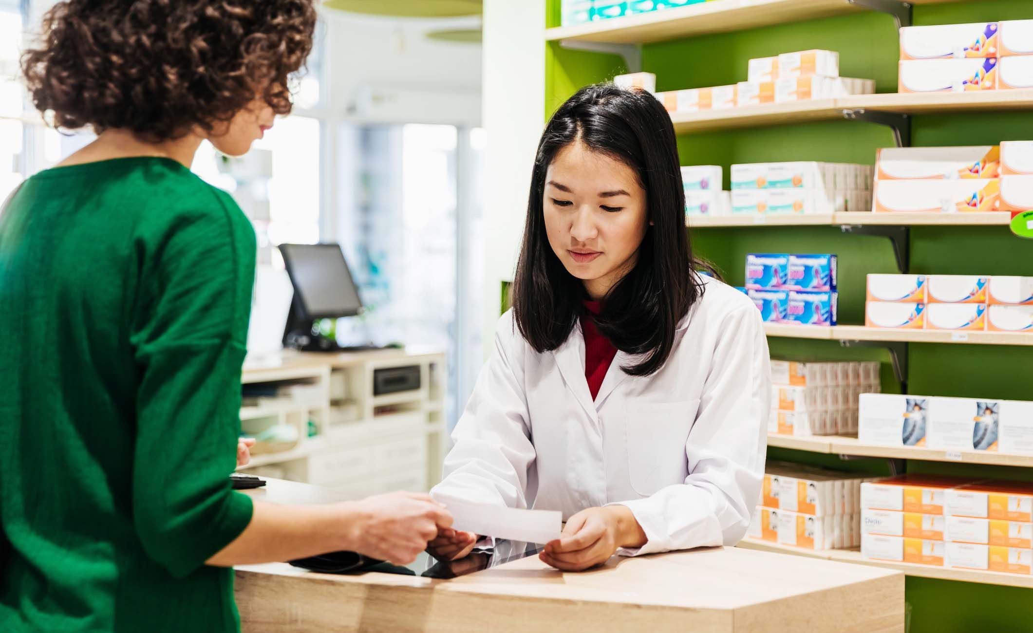 Eine PTA berät eine Kundin im Verkaufsraum einer Apotheke zu einem Medikament.