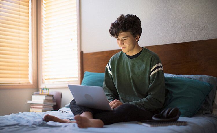 Junge sitzt auf einem Bett mit Laptop auf dem Schoß und beantragt Wohngeld.